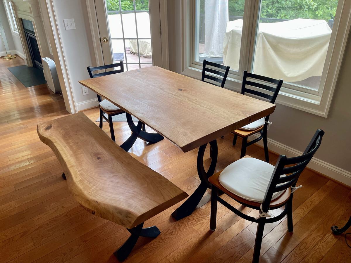 A live-edge slab dining table on black arched steel legs with a matching live-edge bench and ladder-back chairs, in a sunny room with hardwood floors